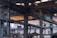 An industrial warehouse scene featuring large, metal structural beams and pipes crisscrossing the space. Overhead, a yellow crane structure with attached cables and pulleys is visible. The interior is dimly lit with natural light filtering in through skylights and windows. The image is filled with industrial textures and machinery components.
