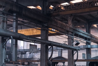 An industrial warehouse scene featuring large, metal structural beams and pipes crisscrossing the space. Overhead, a yellow crane structure with attached cables and pulleys is visible. The interior is dimly lit with natural light filtering in through skylights and windows. The image is filled with industrial textures and machinery components.