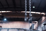 Heavy metal chains with large hooks dangle from the ceiling of an industrial building, with a blurred background showing the high roof structure and metal beams of the interior.