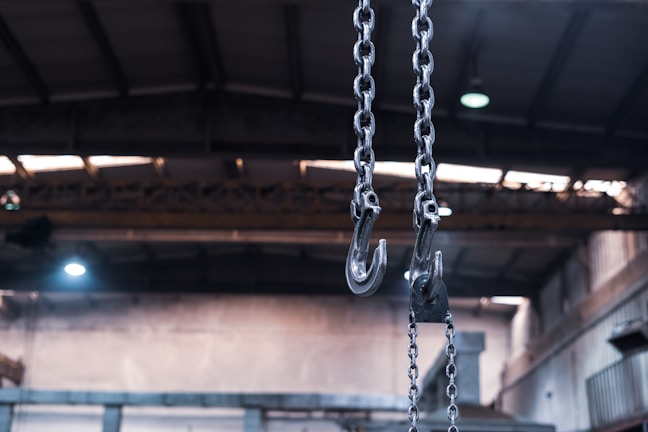 Heavy metal chains with large hooks dangle from the ceiling of an industrial building, with a blurred background showing the high roof structure and metal beams of the interior.