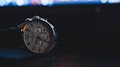 A wristwatch is prominently displayed against a dark background. The watch has a silver casing with a white dial featuring multiple sub-dials and black numbering. The watch hands are luminescent and there is a black leather strap attached. In the background, there are blurred lights, possibly from a computer screen, giving a bokeh effect.