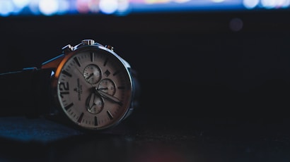 A wristwatch is prominently displayed against a dark background. The watch has a silver casing with a white dial featuring multiple sub-dials and black numbering. The watch hands are luminescent and there is a black leather strap attached. In the background, there are blurred lights, possibly from a computer screen, giving a bokeh effect.