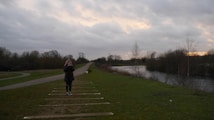 A person stands on a grassy path in a natural setting, holding a camera. The path is flanked by trees and a river runs alongside. The sky is overcast with grey clouds, and there's a hint of sunset hues on the horizon.