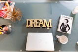 On a glass table, there is a gold decorative object and the word 'DREAM' in bold gold letters. To the left, a collection of fashion magazines and a silver cup with colorful pens can be seen. On the right, there is a book about Chanel featuring a black-and-white image of a woman, and an empty white mug. A closed silver laptop is centered on the table.