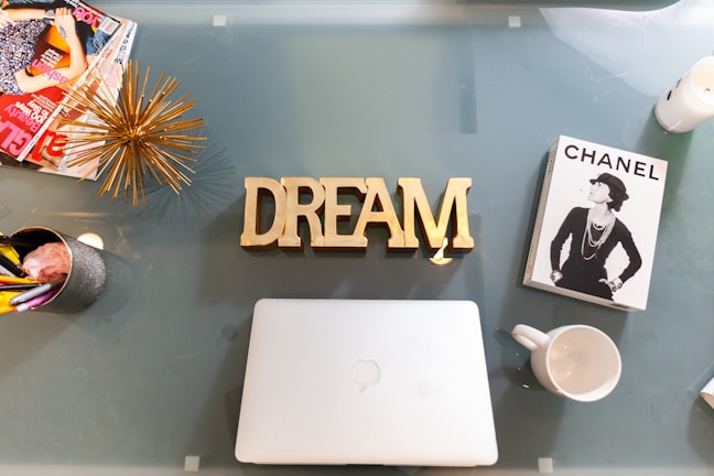 On a glass table, there is a gold decorative object and the word 'DREAM' in bold gold letters. To the left, a collection of fashion magazines and a silver cup with colorful pens can be seen. On the right, there is a book about Chanel featuring a black-and-white image of a woman, and an empty white mug. A closed silver laptop is centered on the table.