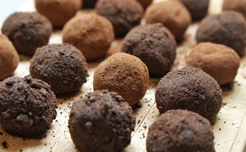 Chocolate truffles are arranged in a close-up view on a wooden surface. Some truffles are coated in cocoa powder while others have a darker, crumbly chocolate coating.