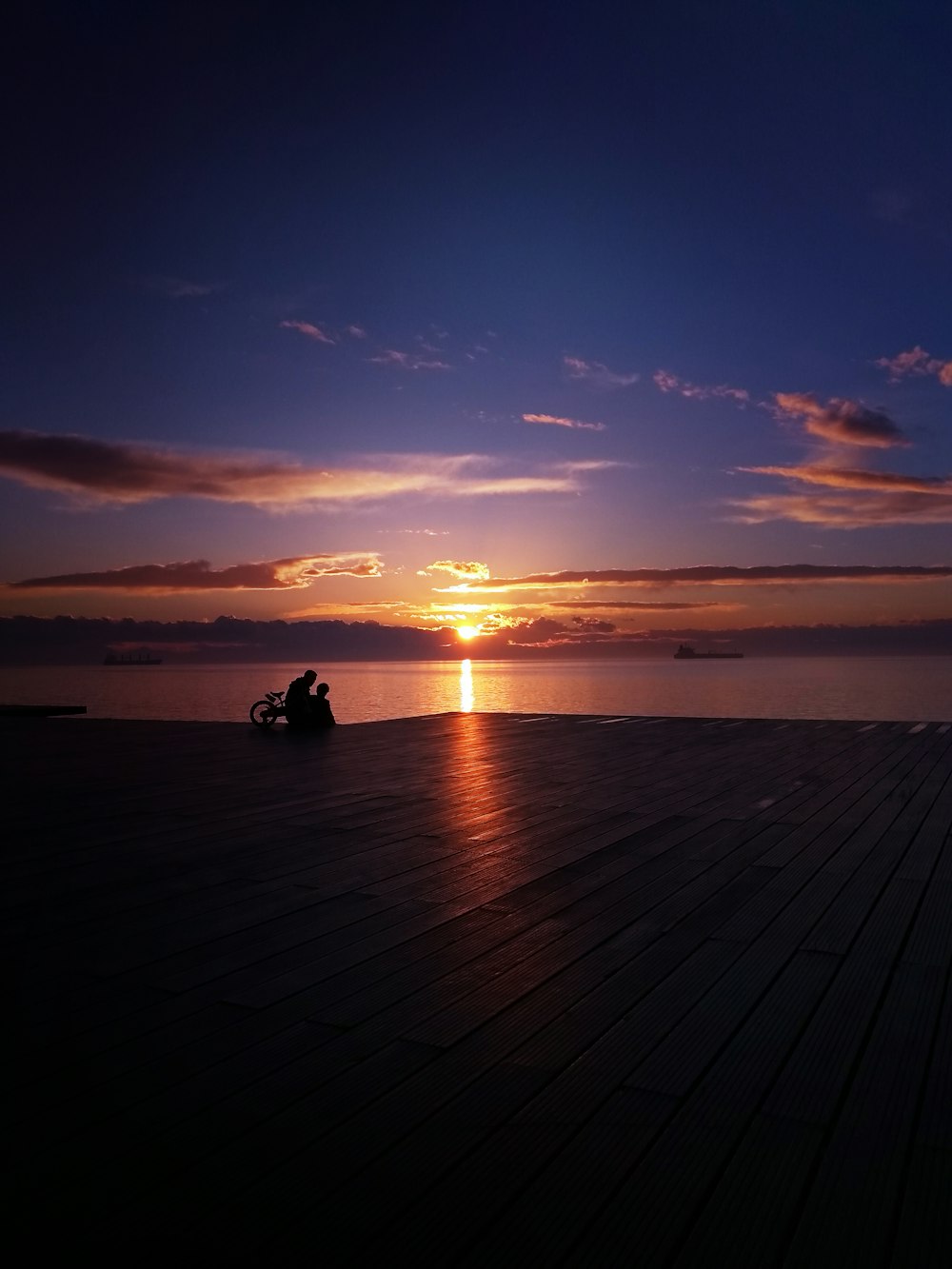 silhouette of 2 people sitting on wooden dock during sunset