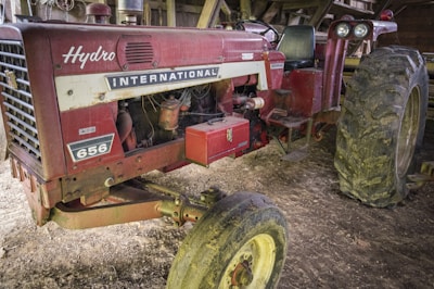 A vintage red tractor with the brand name 'International' and model number '656' displayed on it. The tractor appears well-used, with mud on the large rear tires. It is parked inside a shed or barn with wooden beams visible in the background.