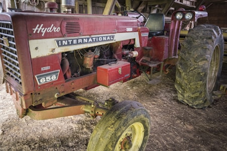 A vintage red tractor with the brand name 'International' and model number '656' displayed on it. The tractor appears well-used, with mud on the large rear tires. It is parked inside a shed or barn with wooden beams visible in the background.