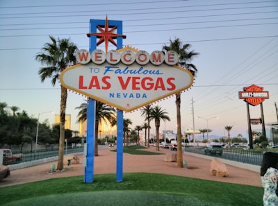A prominent 'Welcome to Fabulous Las Vegas, Nevada' sign is located in the center, surrounded by palm trees and a well-maintained landscaped area. Buildings and distant mountains are visible in the background. A Harley-Davidson sign is seen on the right side.