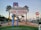 A prominent 'Welcome to Fabulous Las Vegas, Nevada' sign is located in the center, surrounded by palm trees and a well-maintained landscaped area. Buildings and distant mountains are visible in the background. A Harley-Davidson sign is seen on the right side.