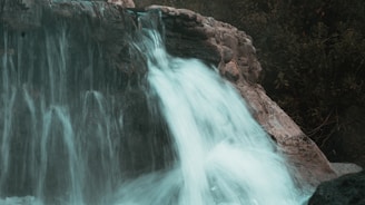 A serene waterfall cascades over rocks surrounded by lush greenery. The water flows smoothly, creating a tranquil and peaceful atmosphere.