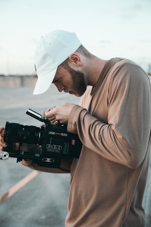 A person wearing a white baseball cap and a beige sweater is closely examining a professional RED RAVEN camera, appearing focused on the viewfinder. The background includes an outdoor setting with neutral, muted tones.