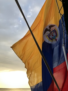 A flag with reencial yellow, blue, and red horizontal stripes with an emblem in the center is seen waving in the wind. The background shows a cloudy sky with the sun setting, casting a warm light over a calm sea.