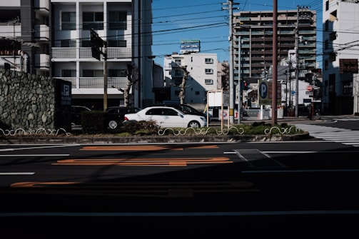 An urban street scene featuring parked cars, residential buildings, and signage indicating a parking area. There are overhead electric wires crisscrossing the view and some traffic lights are visible. A mix of older and newer buildings, with one in the foreground having a stone facade. The street markings are prominently seen on the asphalt.