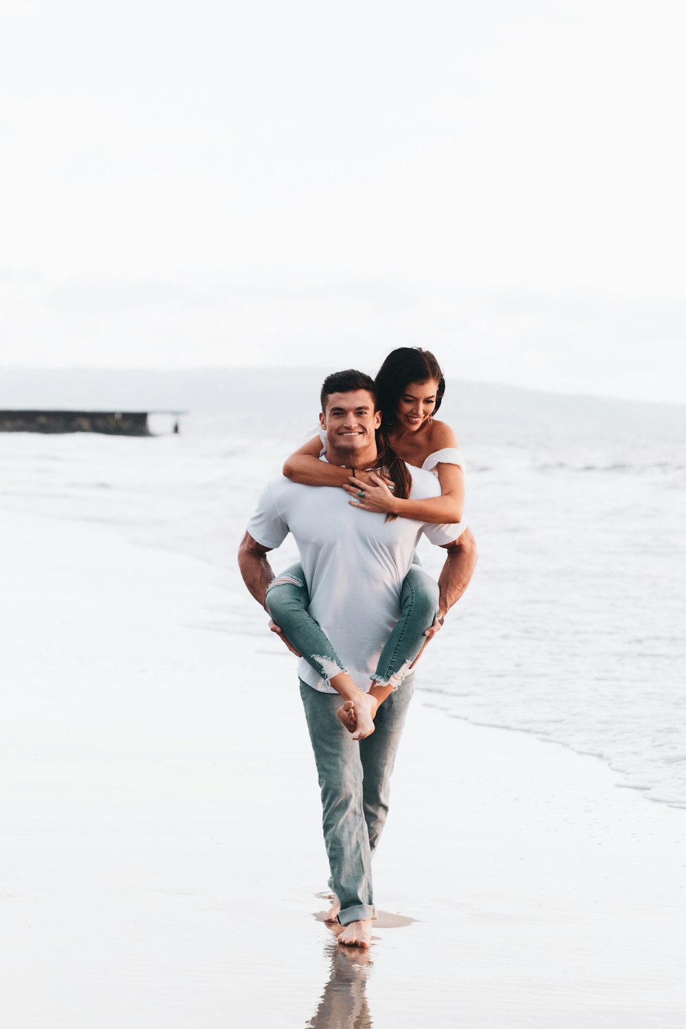 man in blue denim jeans carrying woman in white tank top on beach during daytime