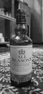 A bottle of All Seasons Golden Collection Reserve Whisky is placed on a patterned surface. The label displays a crest and text describing the whisky as a carefully crafted beverage. The background includes blurred indoor elements that appear to be part of a room.