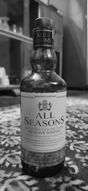 A bottle of All Seasons Golden Collection Reserve Whisky is placed on a patterned surface. The label displays a crest and text describing the whisky as a carefully crafted beverage. The background includes blurred indoor elements that appear to be part of a room.