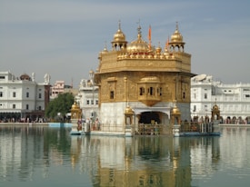 A stunning golden temple stands majestically on a serene body of water, surrounded by an intricate white marble structure. The temple features intricate carvings and a series of golden domes, with a clear reflection visible in the water. Several people are visible around the temple, adding a sense of scale and activity. The sky is a light blue with scattered clouds, providing a peaceful backdrop.