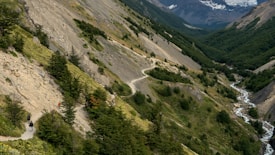 A lush, green valley with a winding path surrounded by dense forests and steep hillsides. The path is being traversed by several hikers. To the right, a stream runs along the base of the hills, adding to the scenic beauty. In the distance, mountains and glaciers are visible under a cloudy sky.