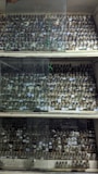A large collection of small glass bottles with cork stoppers arranged on wooden shelves. The shelves are enclosed with glass panels. Each bottle appears to contain a substance, possibly chemicals or samples, with labels on them.