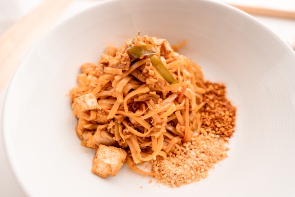 A bowl of Pad Thai noodles garnished with chopped peanuts and crushed chili flakes. The dish features stir-fried noodles, tofu, and appears to be seasoned with traditional Thai spices, presented in a clean, white bowl.