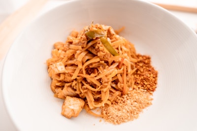 A bowl of Pad Thai noodles garnished with chopped peanuts and crushed chili flakes. The dish features stir-fried noodles, tofu, and appears to be seasoned with traditional Thai spices, presented in a clean, white bowl.
