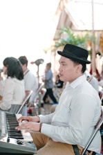 A person wearing a black hat and a white shirt is playing a keyboard in what appears to be an indoor event. Several other people are seated nearby, some facing the opposite direction. The scene has a bright, airy atmosphere with natural light filtering in. In the background, there is blurred greenery and decorative elements.