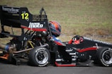 A person wearing a helmet and driving gear sits in a formula race car with visible branding from RMIT University and Team Moto. The car is black with red accents and the number 12 is prominently displayed. The scene is set on a paved road with a grassy background.
