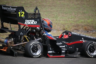 A person wearing a helmet and driving gear sits in a formula race car with visible branding from RMIT University and Team Moto. The car is black with red accents and the number 12 is prominently displayed. The scene is set on a paved road with a grassy background.