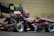 A person wearing a helmet and driving gear sits in a formula race car with visible branding from RMIT University and Team Moto. The car is black with red accents and the number 12 is prominently displayed. The scene is set on a paved road with a grassy background.