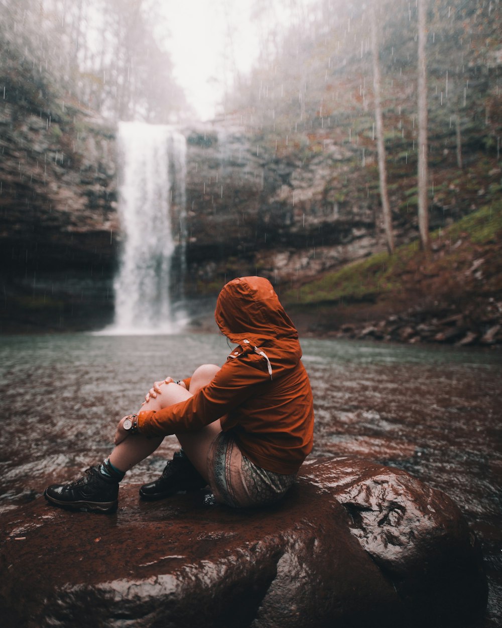 woman in brown hoodie sitting on rock near waterfalls during daytime
