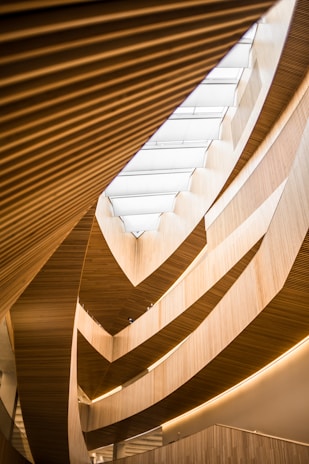 An architectural interior featuring multiple layers of wooden slats and curved structures, which create a flowing, organic pattern. Natural light filters through a large skylight above, highlighting the warm hues of the wood and the gentle curves of the design.