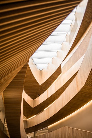 An architectural interior featuring multiple layers of wooden slats and curved structures, which create a flowing, organic pattern. Natural light filters through a large skylight above, highlighting the warm hues of the wood and the gentle curves of the design.