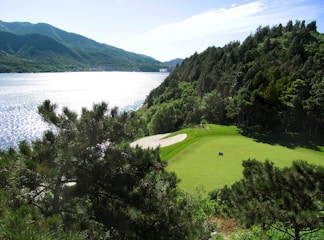 A serene golf course hole is situated near a large, reflective body of water, surrounded by lush, green trees and dramatic hills in the background. A perfectly manicured green includes a flag, and there's a sand trap nearby. The sunlight illuminates the scene, creating sparkling reflections on the water’s surface.