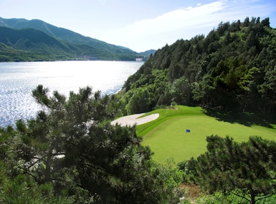 A serene golf course hole is situated near a large, reflective body of water, surrounded by lush, green trees and dramatic hills in the background. A perfectly manicured green includes a flag, and there's a sand trap nearby. The sunlight illuminates the scene, creating sparkling reflections on the water’s surface.