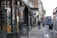 A narrow street lined with shops and a pub, featuring a sign advertising a breakfast special. A double-decker bus is parked along the road, and pedestrians are walking on the sidewalk. The architecture is traditional, with hanging lanterns and flower baskets adding charm to the scene.