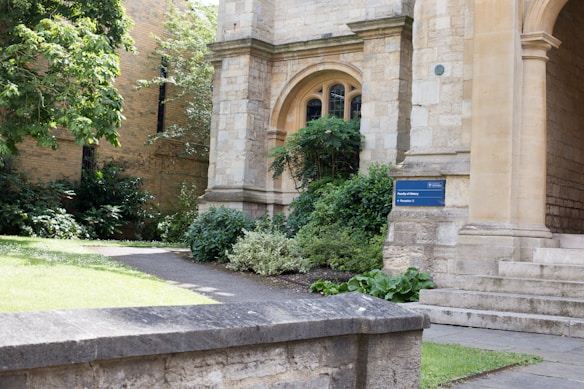 A historical stone building with arched windows and a well-maintained garden in front. The structure features large stones and intricate architectural details. A small path runs next to the building, surrounded by lush greenery and neatly trimmed bushes. A sign with text indicating 'Faculty of History' is mounted on the wall near the entrance.