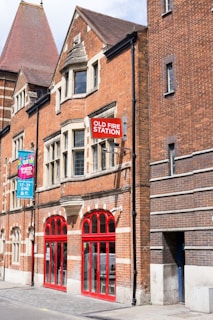 A historic brick building with a prominent red sign reading 'Old Fire Station'. The architecture features arched windows and a mix of red and dark brickwork. A colorful banner hangs on the left, advertising a festival event. The bright red doors and window frames stand out against the brick facade.