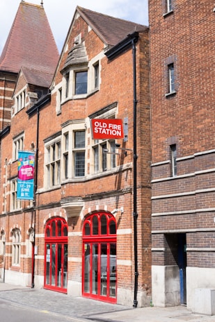 A historic brick building with a prominent red sign reading 'Old Fire Station'. The architecture features arched windows and a mix of red and dark brickwork. A colorful banner hangs on the left, advertising a festival event. The bright red doors and window frames stand out against the brick facade.