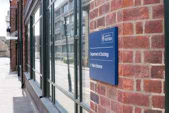 A brick building with a blue sign indicating the location of the Department of Sociology at the University of Oxford. The sign points toward the main entrance, which is in the direction of the glass windows reflecting the opposite street.