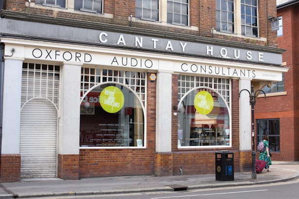 A storefront labeled 'Cantay House' that houses 'Oxford Audio Consultants' with large arched windows. The shop appears to be in an older brick building with white paint surrounding the windows and door. A woman in a green dress is walking on the sidewalk, and a public trash can is visible nearby.