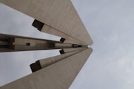 A monumental structure with three prominent vertical slabs rising towards the sky, creating an impression of grandiosity and power. The structure appears to be made of stone or concrete and is set against a cloudy sky, enhancing its monumental feel.