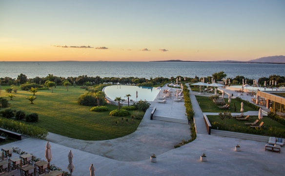 A serene coastal landscape featuring a large, modern infinity pool surrounded by lush greenery and neatly arranged sun loungers with umbrellas. The area is well-maintained with manicured lawns and decorative plants. Beyond the pool, the calm sea stretches into the distance, illuminated by the warm glow of a sunset. The sky is clear with a few scattered clouds, creating a tranquil atmosphere.