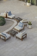 A minimalist outdoor patio setup featuring several cushioned wooden chairs arranged around small square wooden tables. The scene includes potted plants and a round gravel garden area, bordered by verdant shrubs. The tiled flooring is light-colored, enhancing the serene and airy ambiance of the space.
