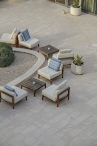 A minimalist outdoor patio setup featuring several cushioned wooden chairs arranged around small square wooden tables. The scene includes potted plants and a round gravel garden area, bordered by verdant shrubs. The tiled flooring is light-colored, enhancing the serene and airy ambiance of the space.