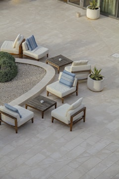 A minimalist outdoor patio setup featuring several cushioned wooden chairs arranged around small square wooden tables. The scene includes potted plants and a round gravel garden area, bordered by verdant shrubs. The tiled flooring is light-colored, enhancing the serene and airy ambiance of the space.