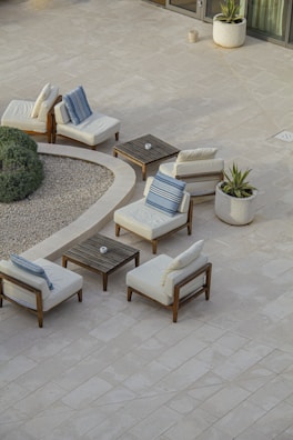 A minimalist outdoor patio setup featuring several cushioned wooden chairs arranged around small square wooden tables. The scene includes potted plants and a round gravel garden area, bordered by verdant shrubs. The tiled flooring is light-colored, enhancing the serene and airy ambiance of the space.