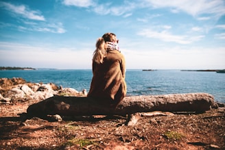 woman in brown sweater sitting on rock near sea during daytime
