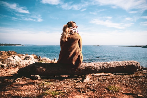 woman in brown sweater sitting on rock near sea during daytime